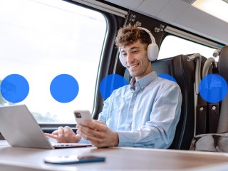 Un hombre joven trabajando sentado en la mesa de un tren con su móvil y su portátil