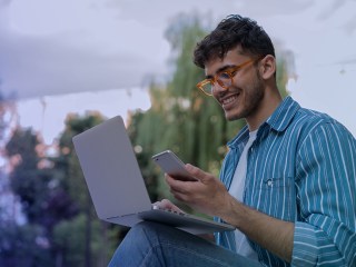 Chico con gafas y pelo corto sonriendo mientras mira su ordenador que sostiene en la mano derecha y su teléfono móvil en la izquierda.