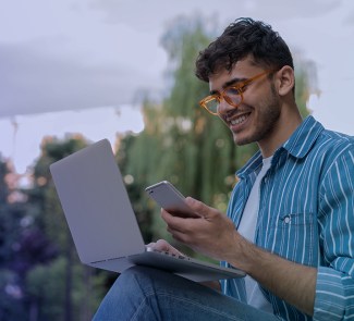 Chico con gafas y pelo corto sonriendo mientras mira su ordenador que sostiene en la mano derecha y su teléfono móvil en la izquierda.